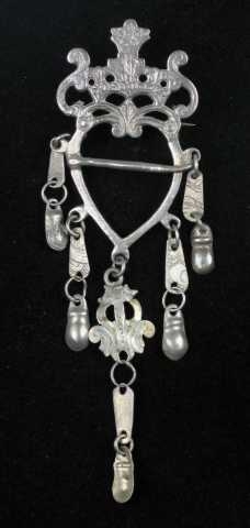 Støpt hjertesprette med dingel. Toppstykket er en gjennombrutt krone flankert av to s-formede ranker. Hele toppstykket har rester etter punkt- og strekdekor. I ett med hjertegrinda er det støpt fem løkkehull der dingelet er festet. Dette består av to eller tre ledd, flate eller drevne små sølvplater forbundet med ringer. Leddene nærmest grinda har rankemotiv. Dingelet som henger fra hjertespissen har tre ledd. Det ene leddet her er større har et gjennombrutt rankemotiv. Tornen er loddet fast tvers over hjertegrinda. På baksida er det festet ei nål med bøyle. Ingen synlige stempel.