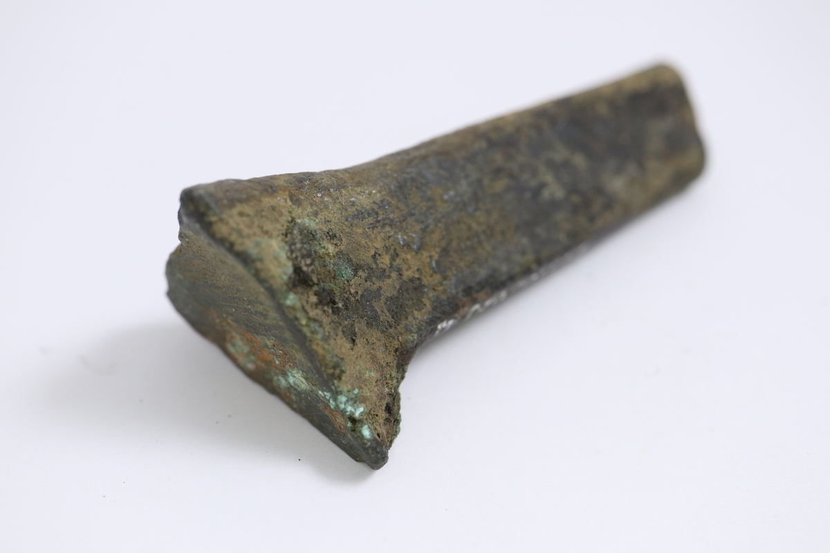 Grytfot av brons med triangulärt tvärsnitt. Breddad upptill mot grytans buk.