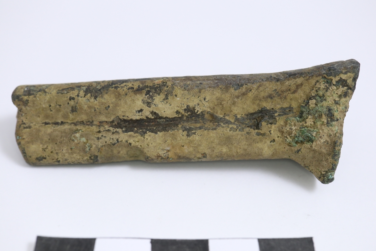 Grytfot av brons med triangulärt tvärsnitt. Breddad upptill mot grytans buk.