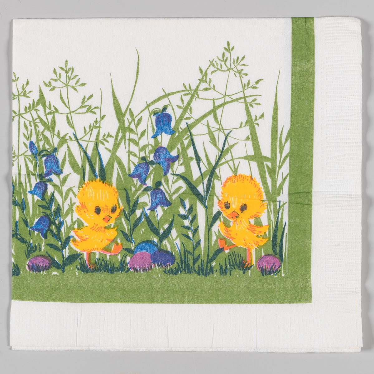 To kylling står mellom høyt gress med klokkeblomster og kulørte påskeegg. Grønn kantstripe.