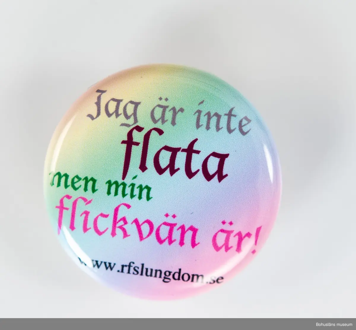 Runt rockmärke med nål för fastsättning på baksidan. 
Bakgrund i regnbågsfärgad gul, grön, blå, rosa med text: Jag är inte flata men min flickvän är! www.rfslungdom.se
[RFSL Ungdom - Ungdomsförbundet för HBTQ-personers rättigheter]
