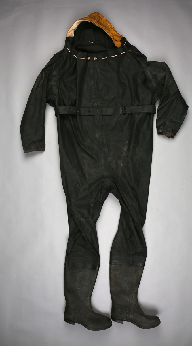 Hel overlevelsesdrakt med støvler, gul hette og belte i livet laget i sort gummi. Åpning i halsen, rundt åpning er det små maljer med tau tredd igjennom. Ved stramming blir drakten tett. Gummisøvlene er integrert i drakten og er relativt store og tunge.