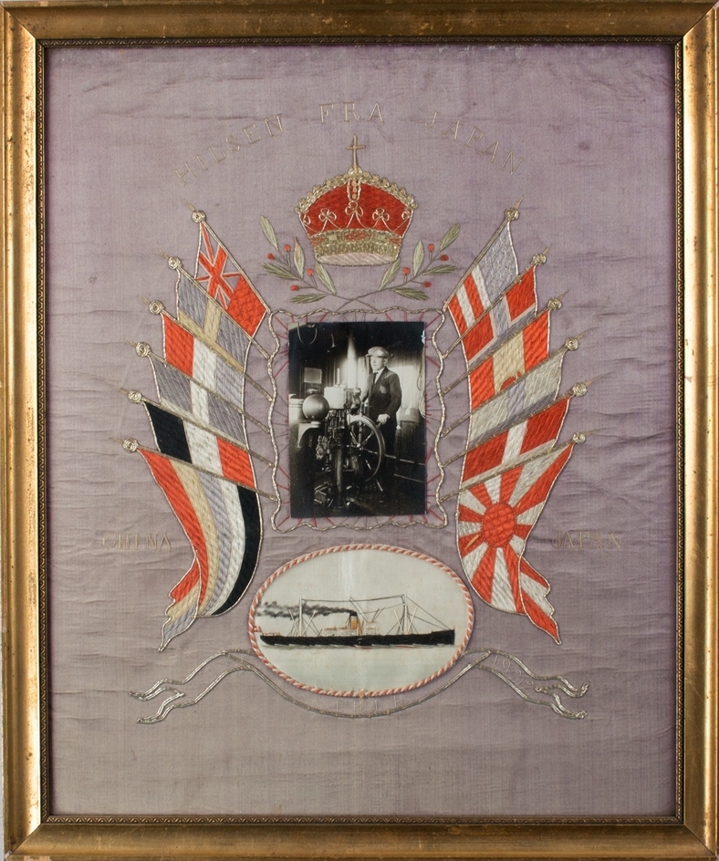 Brodert silkebilde av DS ERLE i stående format. Midt i motivet fotografi av ukjent sjømann til rors, omkranset av seks ulike flagg på hver side og krone øverst. Under et brodert bilde av DS ERLE under fart..