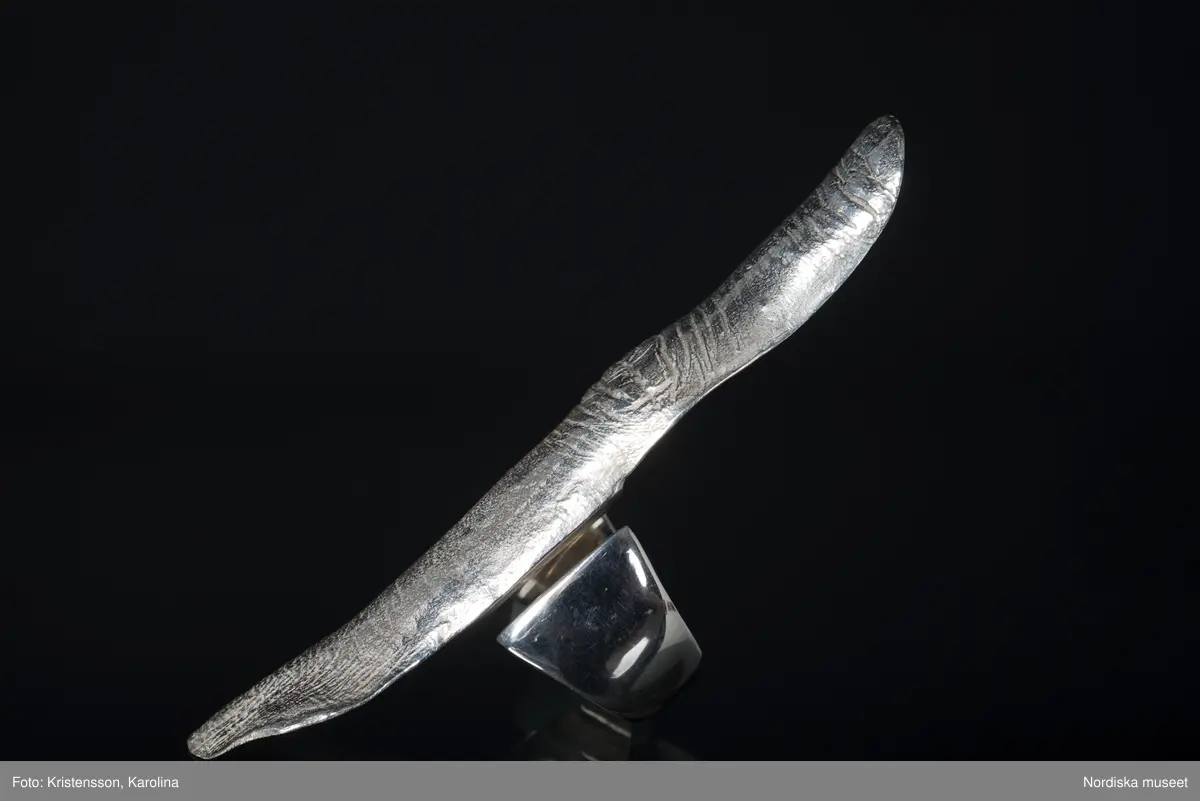 Ring av silver, överdel i form av avlång avgjutning av fingerrygg. Underdel bestående bred ring med slits på ena sidan. Undertill graverad "Cornelia".
/Leif Wallin 2018-02-09