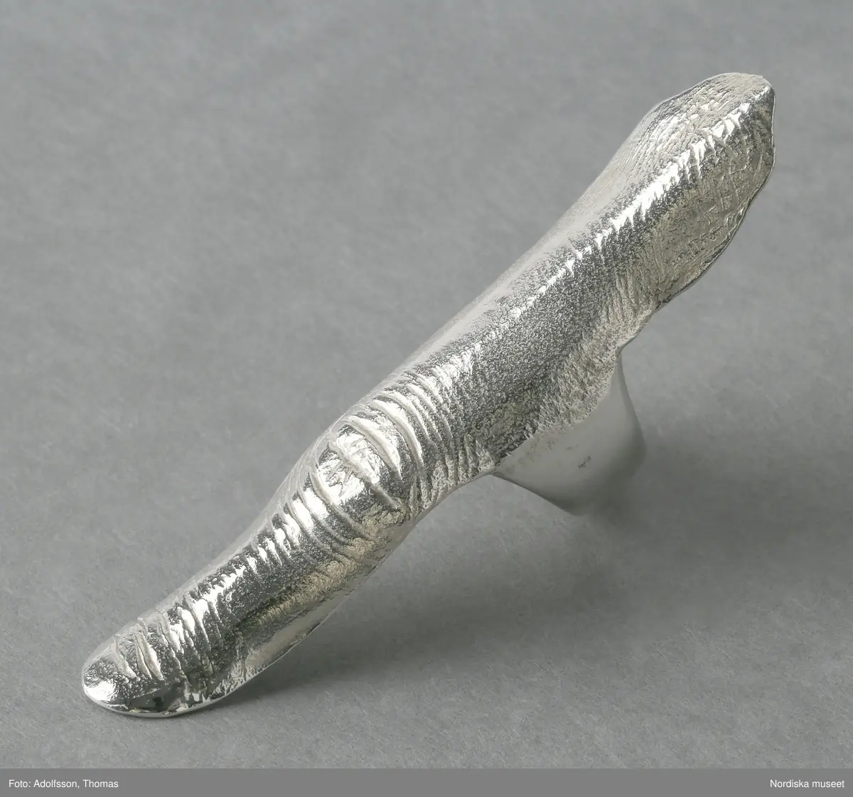 Ring av silver, överdel i form av avlång avgjutning av fingerrygg. Underdel bestående bred ring med slits på ena sidan. Undertill graverad "Cornelia".
/Leif Wallin 2018-02-09