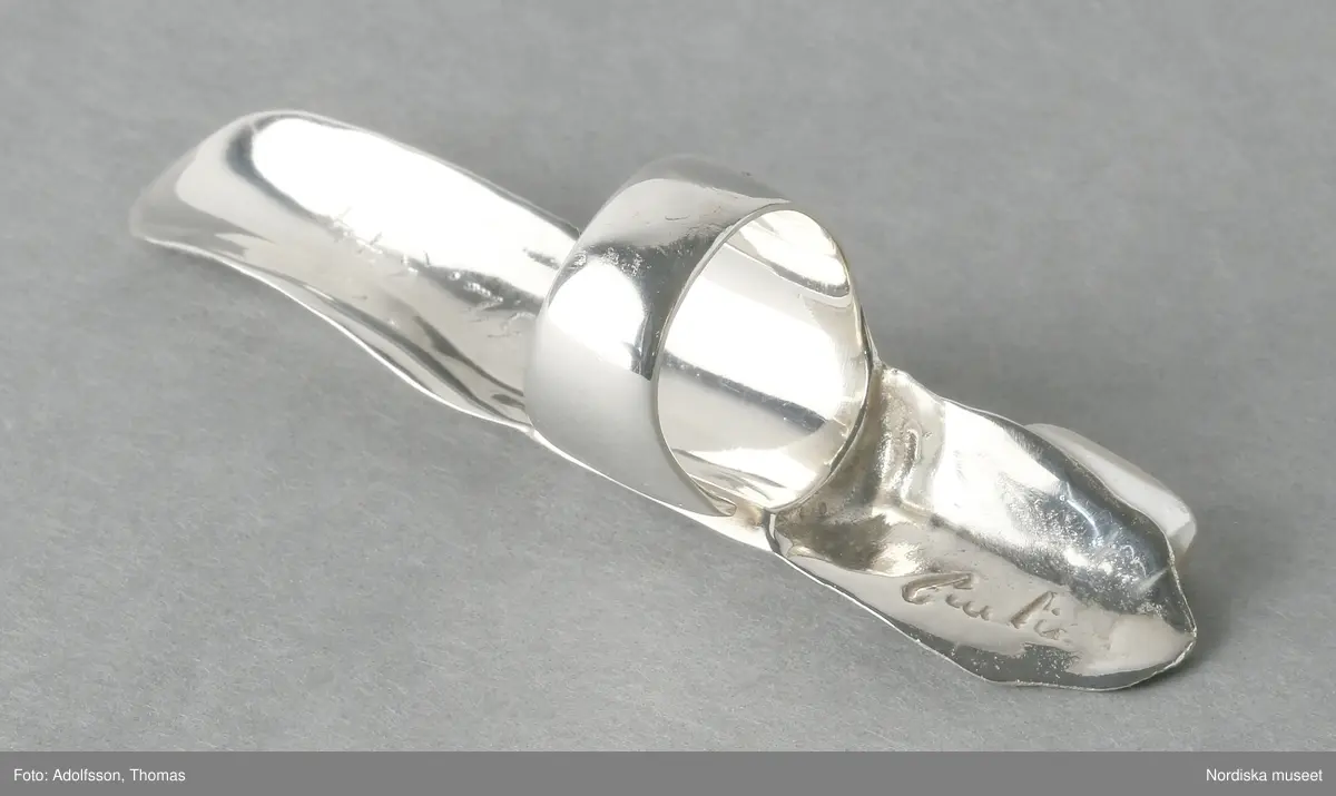 Ring av silver, överdel i form av avlång avgjutning av fingerrygg. Underdel bestående bred ring med slits på ena sidan. Undertill graverad "Cornelia".
/Leif Wallin 2018-02-09
