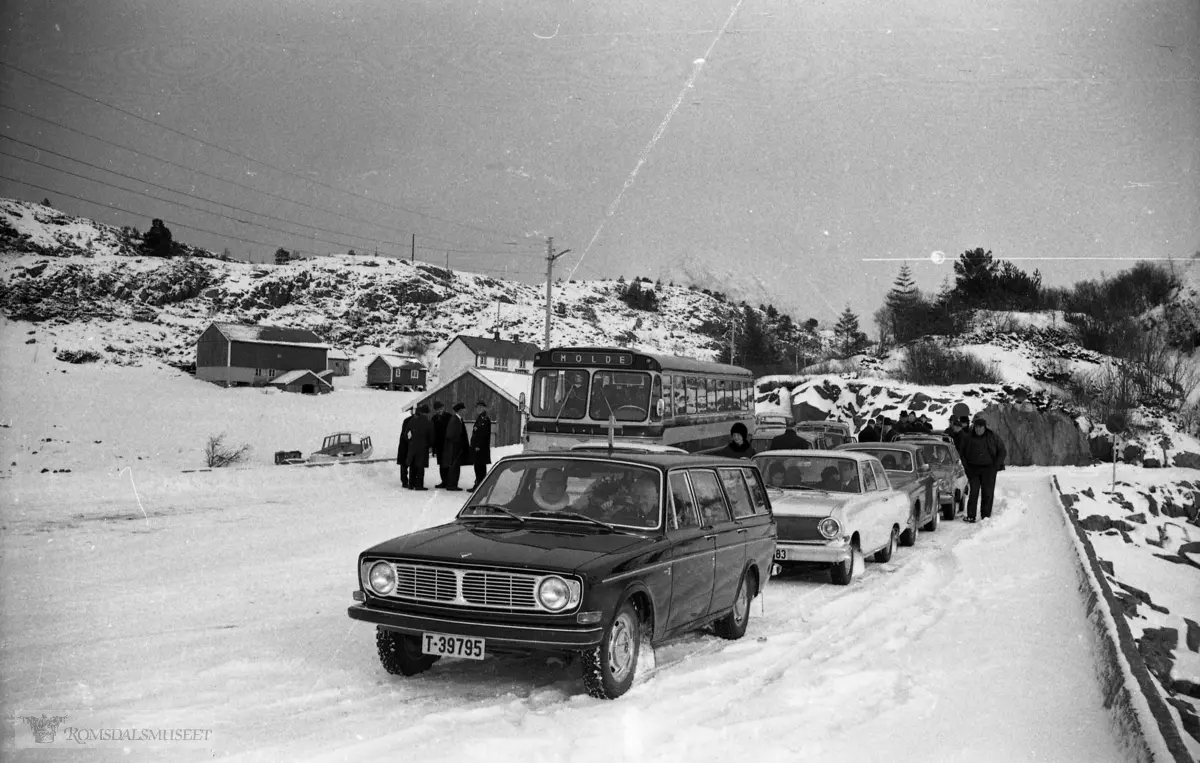 "Mars 1969"."?? mar?"."Kr Heggdal".I bakgrunnen ser vi en buss som er skiltet Molde. Etter fargeskjema å dømme er det en buss fra Aarø og Molde Auto eller Hjelset Auto, lakkert i gult og mørk blå. Etter karosseriet å dømme er dette en buss fra tidsrommet 1966-1968..Disse selskapene hadde ikke ruter over Hollingsholmen, så bussen er antakelig engasjert i turkjøring utenom rute. Bildet er fra 1969 og den gang hadde Aukra Auto og Otrøy Auto ruter over Hollingsholmen. Først i 1974 ble det åpnet eget fergesamband over Julsundet til Otrøya. Det var sambandet Mordalsvågen-Solholmen.