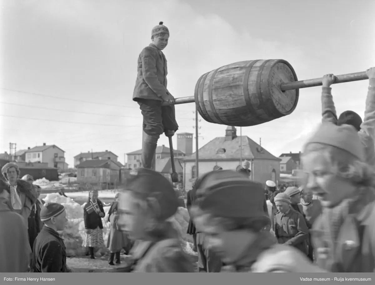 Vadsø 17 mai 1952. Brodtkorps gt. ved "Finnmarksbiblioteket" (hvit murbygning). Folk er samlet til lek og konkurranser, bl.a. speiderjenter der idrettshallen Vadsøhallen ligger idag. En gutt med lue holder en tønne og en annen gutt prøver å klatre opp. Man ser både brakker og gjenreisingsbebyggelse. Bl.a. ser man slangetårnet på brannstasjonen som lå i Idrettsveien. Bygningen ligger der fortsatt, men brannstasjonen er flyttet. Det er ryddet snø.