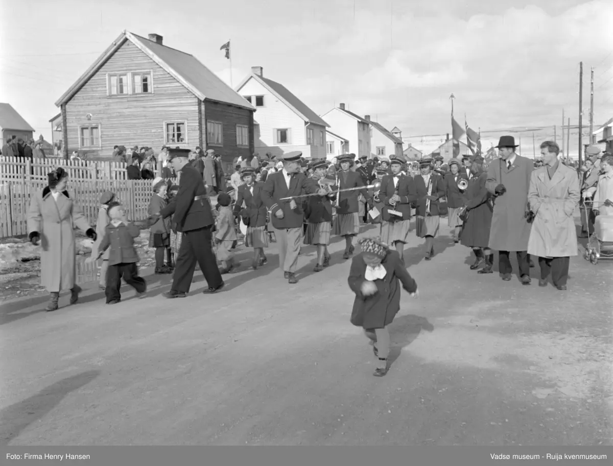 Vadsø 17 mai 1952. 17.mai-toget går gjennom Oscarsgate fra øst mot vest. Ser Melkevarden i bakgrunnen. Jentekorps med dirigent med "taktstokk".  Voksne og barn står langs gata, bl.a. kvinne med barnevogn. Politimann ber folk gi plass for toget.