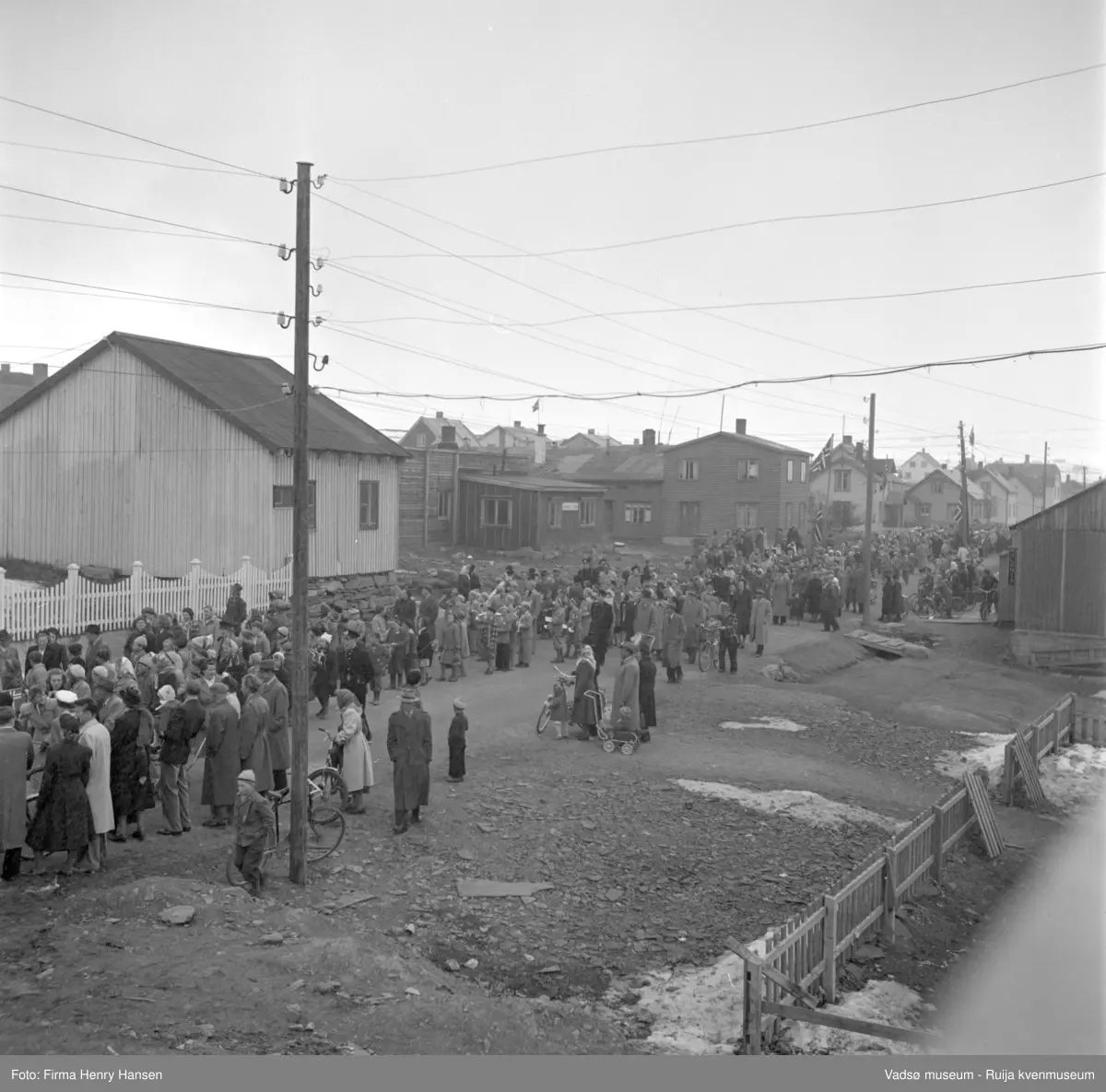 Vadsø 17 mai 1952. 17.maitoget har stanset opp, muligens foran Kysthospitalet som man ser litt av til venstre. Stakittgjerde og det lyse bygget til høyre finner vi igjen på bilder av Kysthospitalet. Sykehuset lå i Oscarsgate, bildet er tatt mot øst. Brakka til høyre for det nærmeste hvite huset har skilt med tekst som ikke kan leses.