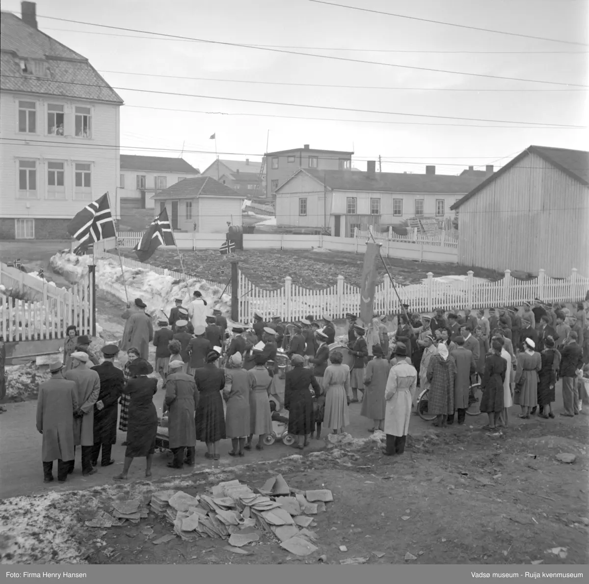 Vadsø 17 mai 1952. barnetoget har tradisjonen tro stoppet opp foran Kysthospitalet i Oscarsgate som da fungerte som byens sykehus. Flaggbærere og jentekorps. Voksne bl.a. med barnevogner og sykkel. Snødunger ligger ennå. I forgrunnen skifer som ble brukt til taktekking. Vi ser flere brakker og ett bolighus