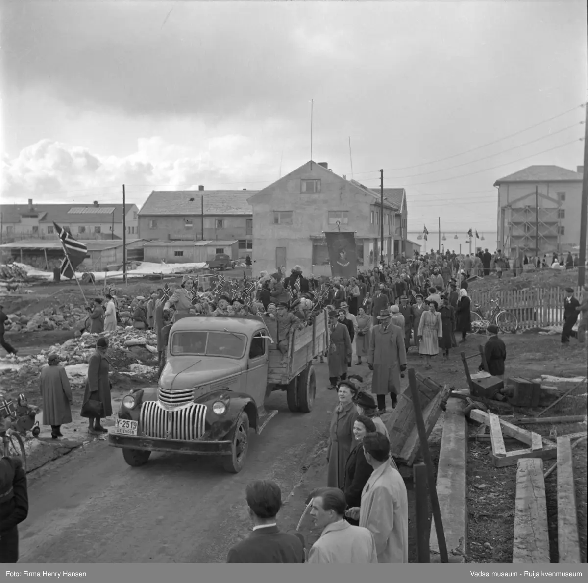 Vadsø 17 mai 1952. Folketog på vei østover i Hvistendalsgate. Vi ser fjorden med fiskebåter i bakgrunnen. Rådhuset med stillas ved torget er nærmest ferdigbygget samt forretningsbygg i typisk "gjenreisingsstil" med bl.a. skifer som taktekking. Fortsatt står brakker og rester etter ruiner. Lastebil med jublende barn med flagg. Lastebilens registreingsnummer er Y-2519 og er en Chevrolet årsmodell 1946-47.

Personer på bild foran til høyre: nr. 2 mann i lys frakk: (N) Mauno, nr. 3 fru Raudio, nr. 4 fru Gallavara, dame midt på bilde til høyre i lys kåpe: Liv Bjørgan Pedersen, til venstre for fru Bjørgan Pedersen - kirketjener Fredriksen og frue,  på lasteplanet på venstre side foran: Ingeborg Grunnes (lotte)