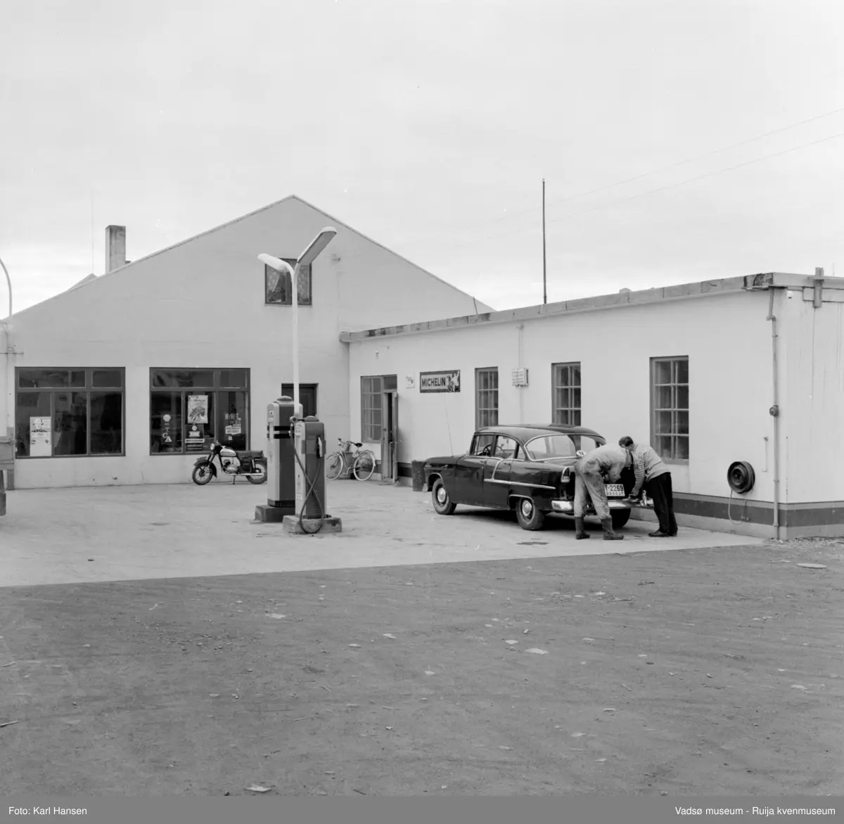 Vadsø sentrum 1959. Shell Bensinstasjon og tankanlegg i Tollbugata. Reklameskilt, moped, drivstoffpumper og en drosje i forkant av bygningen.