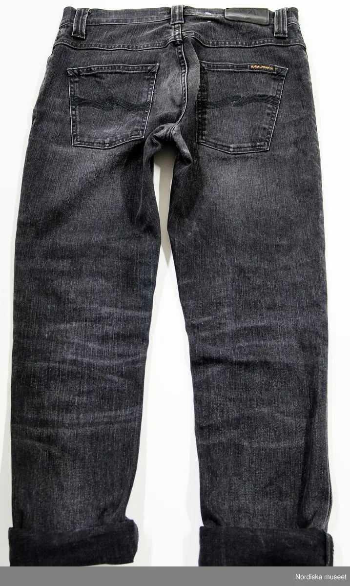 Jeans, av svart tvättad bomull (99%) och Elastan (1%) i kypert. Tyget ska ge karatkär av att vara lite urtvättade och delvis nötta genom att tyget ej är helt svart samt det finns på konstgjord väg innötta veck på tyget på framsidan av låren och baksidan av knäna. Framsidan av knäna är också något "nötta" (lite ljusare mitt på knäna). 
Tyget är töjbart. 
Fem fickor: två framfickor och liten 'myntficka' vid höger framficka, samt två utanpåliggande bakfickor med typisk dekorsöm för Nudies i svart mitt på. Alla fickor förstärkta med metallnitar med text: "Nudie Jeans Co". Gylf fram med 4 metallknappar, varav 3 dolka av gylfslå. Text på knapparna: "Nudie Jeans Co". 5 hällor i linningen. Höger bak på utanpå linningen svart etikett i läder (?) med pressat "N / Nudie Jeans Co.". Svart mindre tygetikett fastsydd på insidan av linningen bak samt på höger bakficka med text "Nudie Jeans o Co". 
Rester av vit tråd (efter pappersetikett som satt på jeansen i butik) bredvid läderetiketten. 
På insidan fickpåsar fram av grått bomullstyg, På den högra fäst etikett med produkt- och skötselinformation samt etikett med storleksangivelse, och på den vänstra tryckt text direkt på fickpåsen (se nedan). 
Jeansen ben är uppvikta, vilket är så som brukaren alltid har använt dem. Uppviket är ca 4,5 cm brett uppvikt 5 "varv". 

Text på etikett på höger fickpåse: "Nudie Jeans Co / www.nudiejeans.com / Nudie jeans marketing AB / Västra Hamngatan 5 / SE-411 17 Göteborg"..."99% organic cotton"...1% Elastande"... tvättsymboler för 40 grader, tål ej klorblekning, ej torktumling (?), strykjärn mellantemperatur (två prickar) Tål inte starkare tvättvätskor än perkloretylen. "...Sash inside out with similar colours / Don not use chlorine bleach / Please stretch inseam while damp."..."May Dry- or Wet bleed / contains non-textile parts of animal. / Made in Italy." samma text på franska och kinesiska (eller japanska). På storleksetiketten: "W[aist] 29 L[ength] 32. / Nj1001353 [produktnummer] / GRIM TIM [Modellnamn] / BLACK HAZE [färg] / 99% ORGANIC COTTON / 1% ea."
Text på vänster fickpåse: "ORGANIC / THE COTTON USED IN THE MAKING OF THIS GARMENT IS ORGANICALLY GROWN AND HARVESTED. / 100% ORGANIC COTTON / WE LOVE JEANS, a passion we share with anyone who mourns a worn-out pair like the passing of a close friend.No fabric ages quite as beautifully as denim; the longer you wear your jeans, the more character and attitude they acquire.You shape them with your lifestyle, and they become like a second skin. Jeans share the same soul and attitude found in the rock’n’roll scene — they’re both part of the same culture.
We’ll always stay true to jeans. We’re not looking for fly-by-night fads; our inspiration is found far from the world of glamour and catwalks. We don’t just offer jeans.We offer a way of thinking, a concept, and an undying passion, fuelled by the traditions of denim and the characteristics of the fabric itself."
/Leif Wallin 2017-08-15