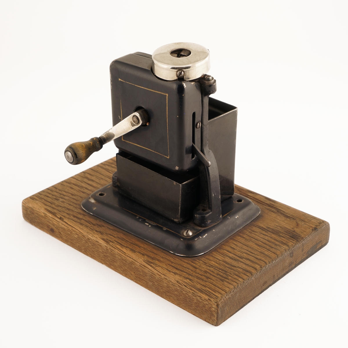 Blyantspisseapparat av  sortlakkert metall montert på treplate. Dreiet håndtak i tre.