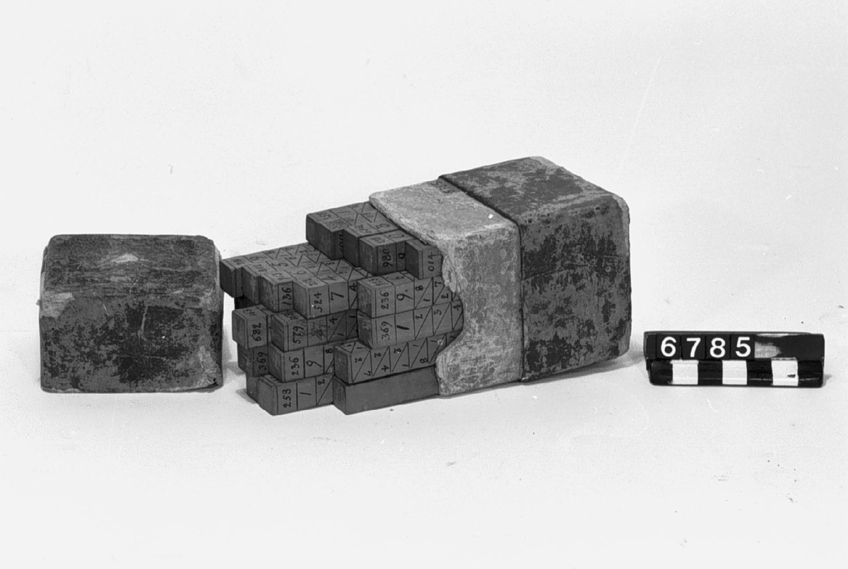 Napiers räknestavar för multiplikation av intill åttasiffriga tal med alla siffror av samma slag. Stavarnas diameter 13 x 13 mm. Komplett med 25 stavar i fodral av papp. Denna serie liknar mest den i katalogen "Mathematics I. Calculating machines and instruments", London 1926 under n:o 7, sid 11 upptagna, men samtliga multiplicandstavar är upptill på alla sidor försedda med siffror, som hänvisa till vilka siffror-multiplar, som befinner sig på övriga sidor. Dessutom finns kvadranterna och kuberna på talen 1-9. 25 st. neperska räknestavar (av trä med siffror i tusch) enligt John Napier (f. 1550, d. 1617, skrev vanligen Neper), för multiplikation av 8-11-siffriga tal med alla siffror av samma slag eller högst 24-siffriga tal. En stav med sifforna 1-9 i var sin vågrät rad på endast en sida bör vid räkning läggas till vänster om stavarna för att undvika förväxling av multipelrad vid produktens avläsning. På motsatta sidan av femte staven fr. v. på fotot finns kvadrater för talen 1-9. Likaså finns en stav med kuber för talen 1-9 på en sida. Stavarnas längd: 90 mm, tvärsnitt 13 x 13 mm. Den i katalog fr. Science Museum uppgivna räknevalsen saknar hänvisning upptill på varje stav till de på dennas övriga sidor befintliga siffrorna med multiplar. Antalet stavsidor med multiplar för vardera talen 0-9: 0  1  2  3  4  5  6  7  8  9 så att multiplikanden kan bestå av högst 10 8  9 10  8 10  10 9 11 11 = 94 sidor 8-11 siffror av samma slag.