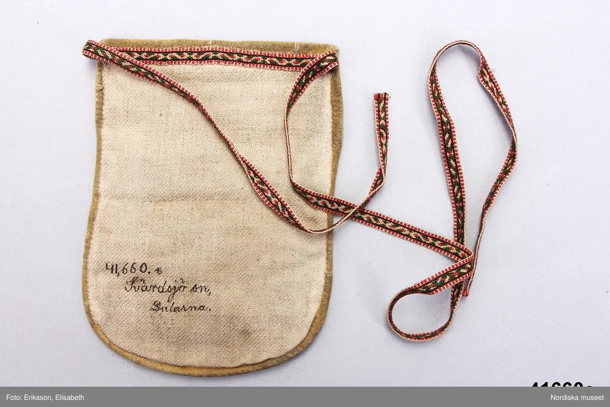 e. Kjolväska av mörkt blåsvart kläde.  kantad med ljust sämskskinn, på fickan applikationsbroderi med rött och gult kläde samt grön vadmal med tenntrådskonturer i läggsöm. Fickan insida av linnelärft, bakstycke av oblekt linnekypert.  Påsytt smalt midjeband av mönstrat halvylleband i vitt lingarn  med plockat mönster i grönt och rosa ullgarn. Avsett att knytas på vänster sida. 
I mycket gott skick
/Berit Eldvik 2012-02-14