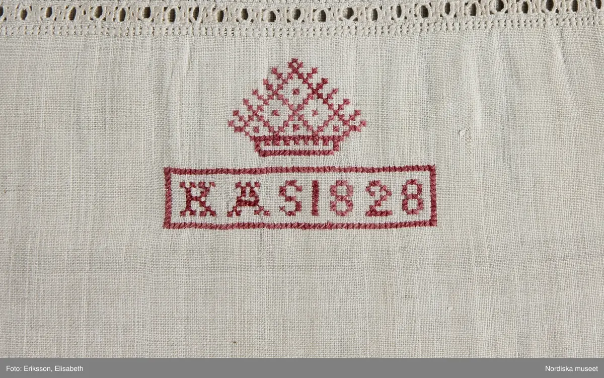 Skjorta av vit linnelärft, ett framstycke och ett bakstycke med halsspjäll som går ända ut mot ärmsömmen. Rynkad runt halsen mot hög uppstående krage kantad med knypplad 3 cm bred linnespets av tät skånsk typ. På halva kragbredden broderad bård i vitt och ljusgrått lingarn i tät utdragssöm av skånsk typ. Halssprund 25 cm långt med ändstopp av en bit s.k. vävd spets. I kragsömmen 2 stora knapphål för länkknapp se litt b.  Runt sprundet och i en kvadrat nedanför rikt broderi i utskårssöm och utdragssöm  samt något plattsöm och hopdragssöm. med vitt lingarn. Nedanför broderiet märkt K A S 1828 under "krona" med rosa bomullsgarn i korssöm. Vidsydd vid ärm med ärmspjäll, nedtill rynkad mot 3 cm bred ärmlinning med stickningar, knutar och enkel utdragssöm samt kantad med lätt rynkad knypplad spets av samma sort som på kragen, ärmsprund med 2 motstående knapphål.
En mycket typisk  brudgumsskjorta av rikaste slag från Ingelstads hd. Broderi och sömmar mycket välgjorda.
/berit Eldvik 2010-10-15
