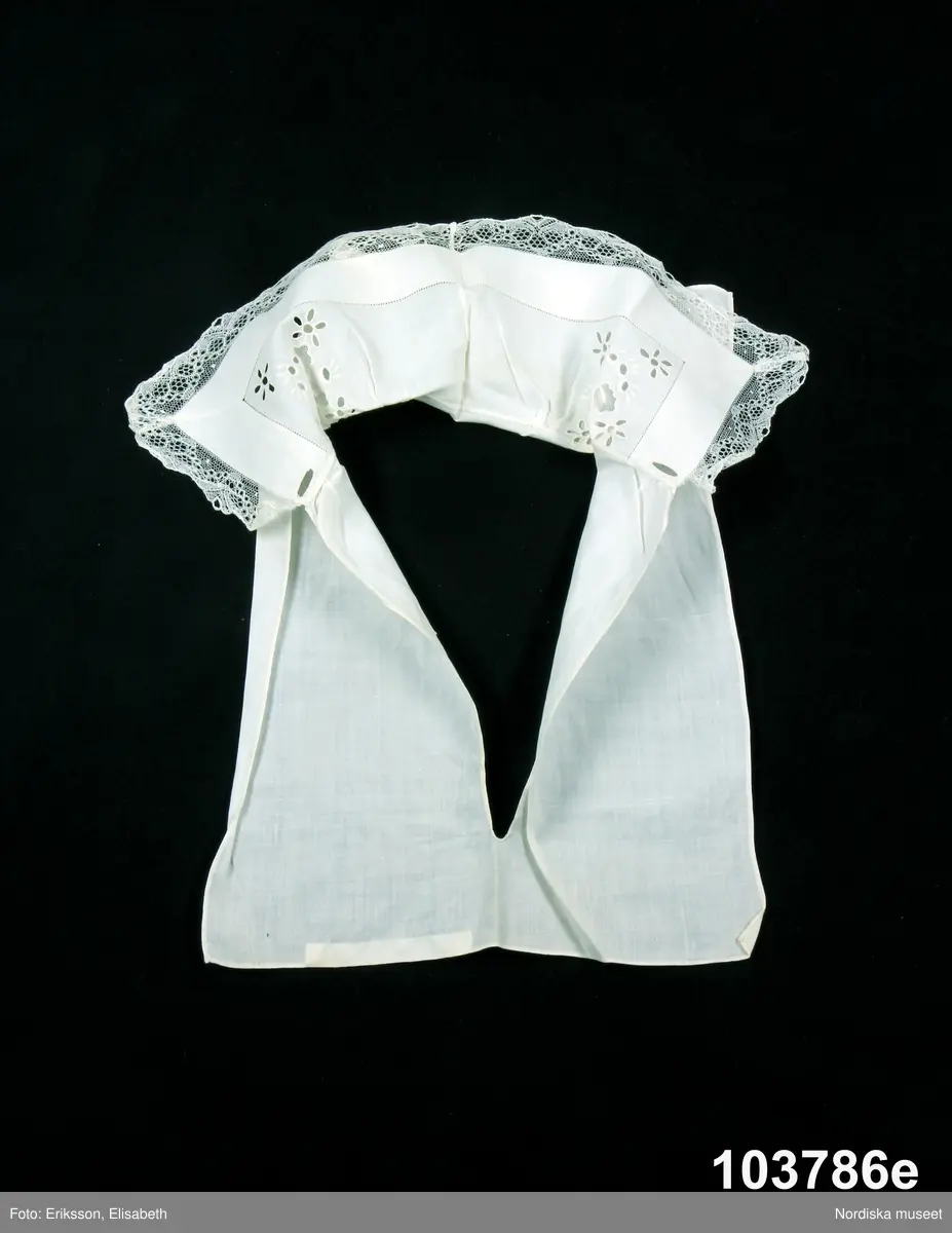 e: Krage. Längd: 47,5, cm - totallängd, bröststycke. Bredd: 33 cm - bröststycke. Höjd: 14 cm - kraghöjd, inkl spets. Material: Textil, bomull. Lingarn - spets. Bomullsgarn - broderi. Teknik: Tuskaft. Vitbroderi, Knyppling. Handsömnad.
Krage i tuskaftat vitt bomullstyg. Rektangulär duk, uppklippt halshål med fastsydd krage, 14 cm hög. Kragen har 3,5 cm bred fåll, enkel utdragssöm i kanten. Vitbroderi  i framkant,  plattsöm och kantsydda hål med infällt tyll, två sydda knapphål. Handknypplad spets fastsydd på långsida och korstsidor, 2,5 cm bred. Öppning fram, 24 cm långt sprund. Kragen är handsydd.
Lästips: Svensson Sigfrid, (1935). Skånes Folkdräkter, en dräkthistorisk undersökning 1500 - 1900. Stockholm: Nordiska Museets Handlingar: 3.
/Inga-Lill Eliasson 2008-02-19
