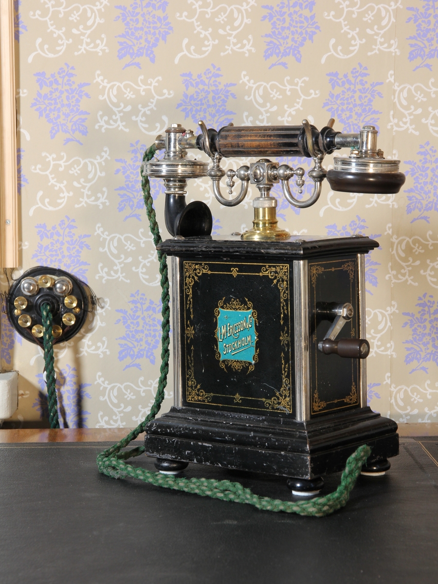 Telefon, bordsapparat med lur. Modell A 381 Jubileumsapparat (framtagen till L M Ericssons 25-årsjubileum 1901). Stående rektangulär med klyka av vitmetall. Svartmålad med dekor och text bland annat i guldfärg. Vev. Väggplint och grön sladd.