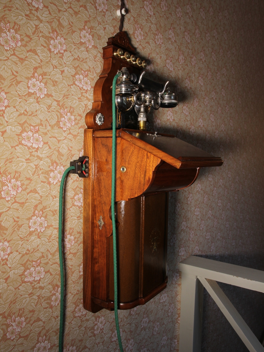 Telefon, väggapparat. Stående rektangulär med snedställd skrivplatta av brunbetsat trä. Klyka av vitmetall, lur och grönt telefonsnöre, Till höger vev. Kompletterad.