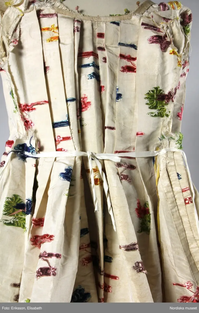 Brudklänning (robe), av broscherat, vitbottnat kinesiskt siden. Nygjord framvåd av fint vitt bomullstyg med kjolvolang nedtill av robens tyg. Två lösa garneringsbårder.