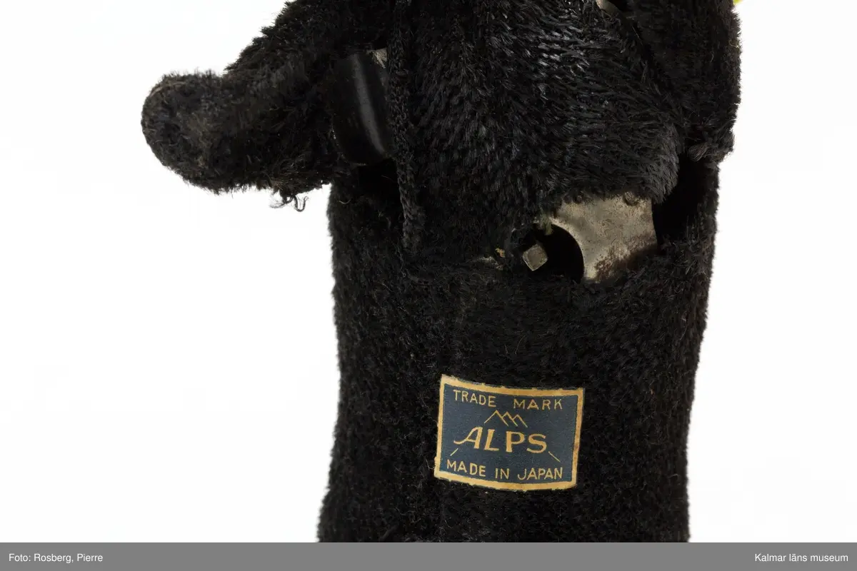 KLM 32004 Hund, leksak, metall, textil och plast ev. celluloid. Mekanisk leksakshund, svart Labrador-liknande som har ett köttben i munnen. Hunden har ett mässingsfärgat halsband. Hunden är gjord av metall med svart plysch utanpå som päls och ögon av plast. Uppdragningsmekanik med nyckel på undersidan, nyckel saknas, så att hunden kunde gå, röra på huvudet och vifta på svansen. Etikett på undersidan med texten: Trade Mark ALPS. Made in Japan.