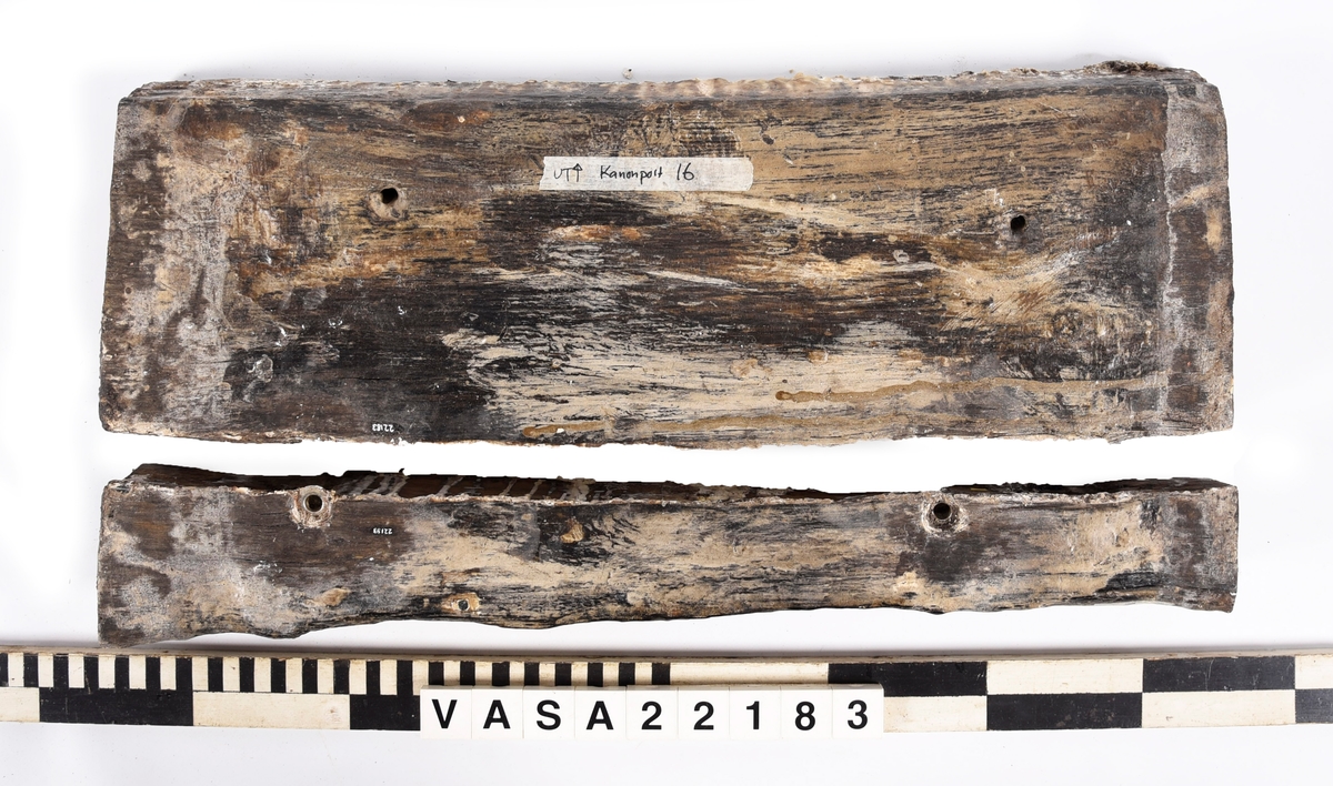 Tröskel till kanonport 16. Paralellogramformad på ändor, fyrkantig på fram och baksidor. 2 klart original spikhål, 6 andra med moderna scruv-spår. 
Splittrad längs en långkant. Kraftig vittrad på alla ytor utomför ändor. Järn impregnering rund alla spikhål på ena yta. Tjock fläckor PEG.