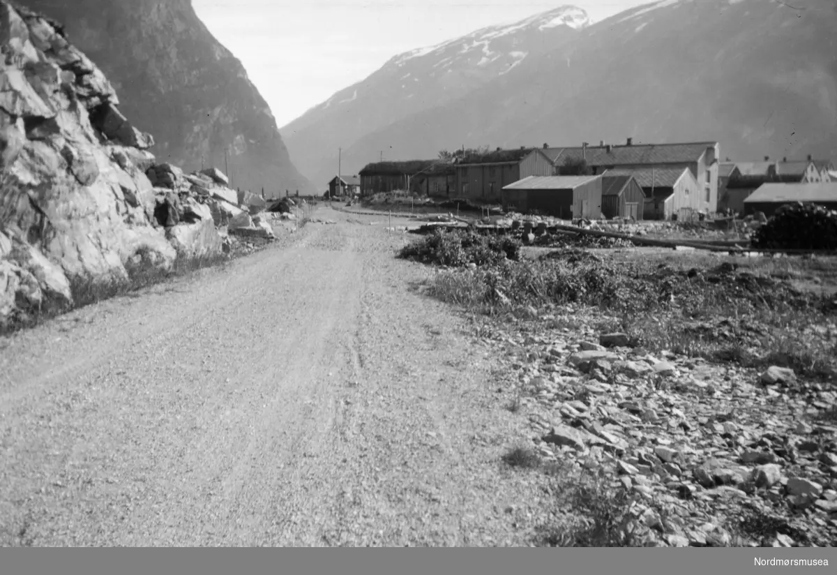 Foto fra en landevei, men en del bebyggelse på høyre side. Det er usikkert hvor, men kan muligens være fra Nesset kommune hvor den som trolig er fotograf (Georg Sverdrup) ofte var på besøk. Fra Sverdrupsamlingen ved Nordmøre museums fotosamlinger.