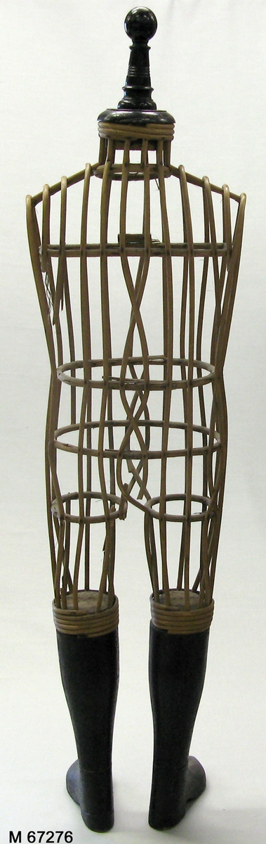 Provdocka av rotting med snidade svartmålade underben/stövlar av snidat trä och svartmålad svarvad avslutning i halsen. Rottingen formad och fastspikad genom vertikala cirklar som håller ut de kroppsformade horisontella rottingbitarna.