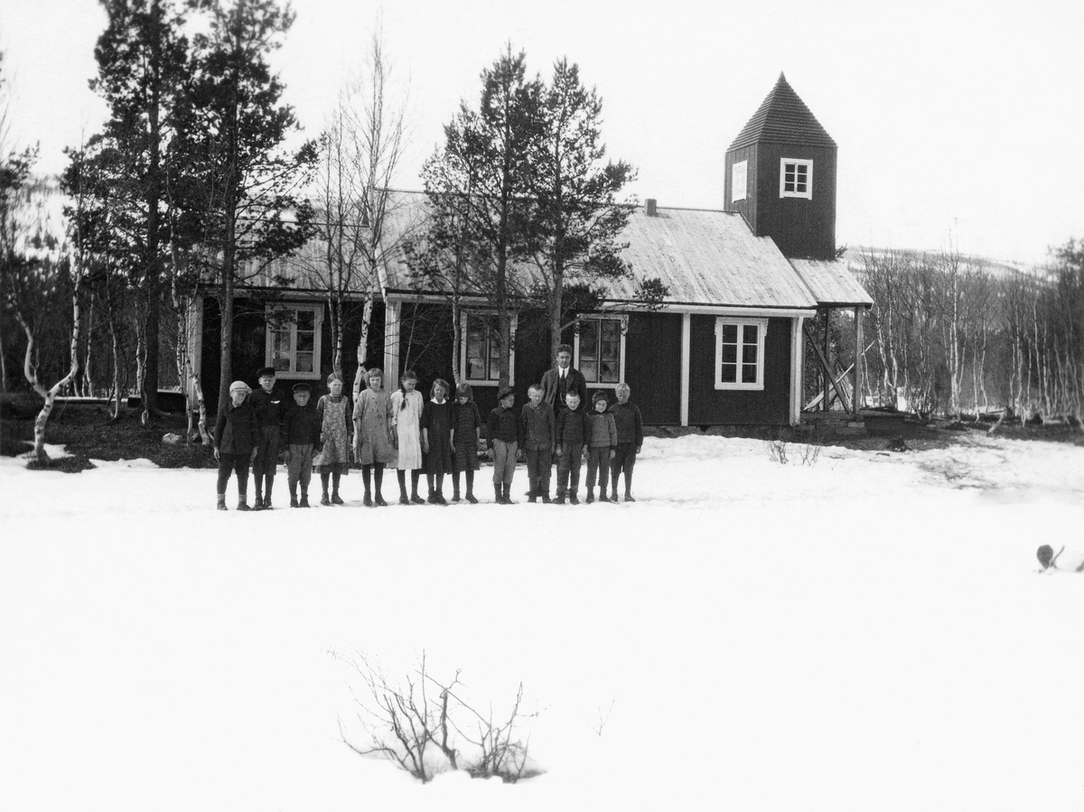 Trallerud skole i Susendalen.  Fotografiet viser tretten elever og en lærer framfor skolebygningen, som med et tårnoppbygg over inngangen på gavlveggen har et visst kapell-preg.  Skolen ligger i fjellskog med bjørkeskog ispedd ei og anna furu.  