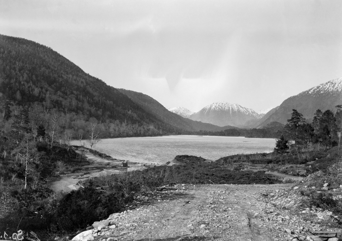 Utsikt over Friskogen i Lyseklosterkomplekset, sett fra sørenden av Rødlivatnet.  Fotografiet er tatt fra en veg mot et vatn med avløp til en liten bekk nederst til venstre i bildet.  Omkring vatnet vokser ei blanding av furu- og bjørkeskog.  Bakenfor Rødlivatnet (i nord) ruver et par markante fjell med snø på toppene. 