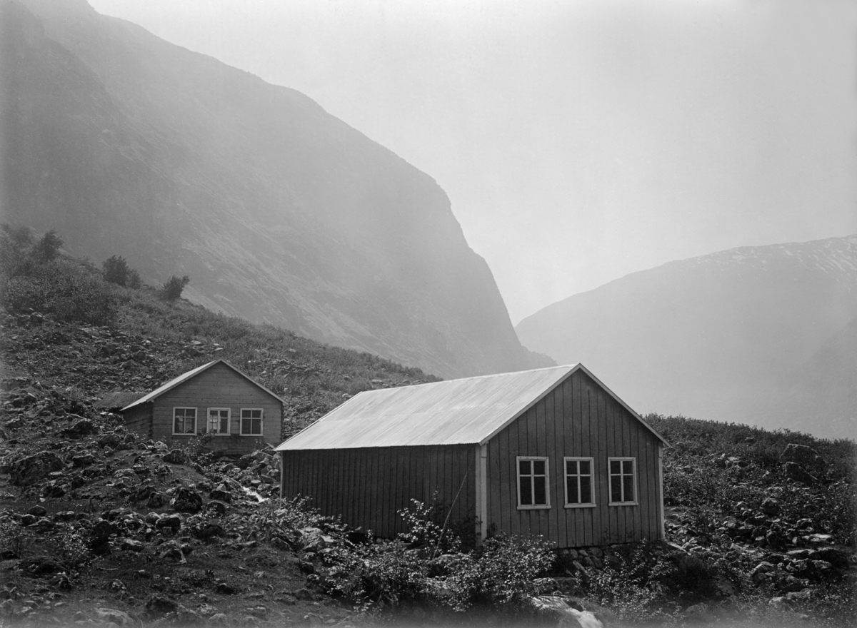 To utklekningshus for settefisk på Hauge i Lærdal (cirka 4 kilometer fra Lærdalsøyri).  Fotografiet viser to klekkerihus, oppført i bindingsverk med vertikal over- og underliggerkledning og saltak, tilsynelatende bølgeblikktekket.  Begge bygningene har tre krysspostvinduer på gavlveggen som vender mot fotografen.  På bakken skimter vi også den lille bekken med vann som rislet gjennom utklekkingsrennene.  Fiskesdammene der fisken fikk vokse seg større og mer robust lå på sletta like nedenfor de avbildete bygningene, jfr. KAS.123.  I bakgrunnen på dette fotografiet skimter vi høye, bratte vestlandsfjell.  

Dette er en del av et fiskeklekkeri- og oppdrettsanlegg som statens fikskeriinspektør fikk bygd i samarbeid med grunneier Anders Jensson Hauge (1857-1938) i 1899, som ledd i et nasjonalt prosjekt for å «forbedre Norges Laxe- og Ferskvannsfiskerier».  Rundt 1900 skal det ha vært om lag 20 slike anlegg i Norge.  Klekkeriet på Hauge ble nedlagt i 1949.