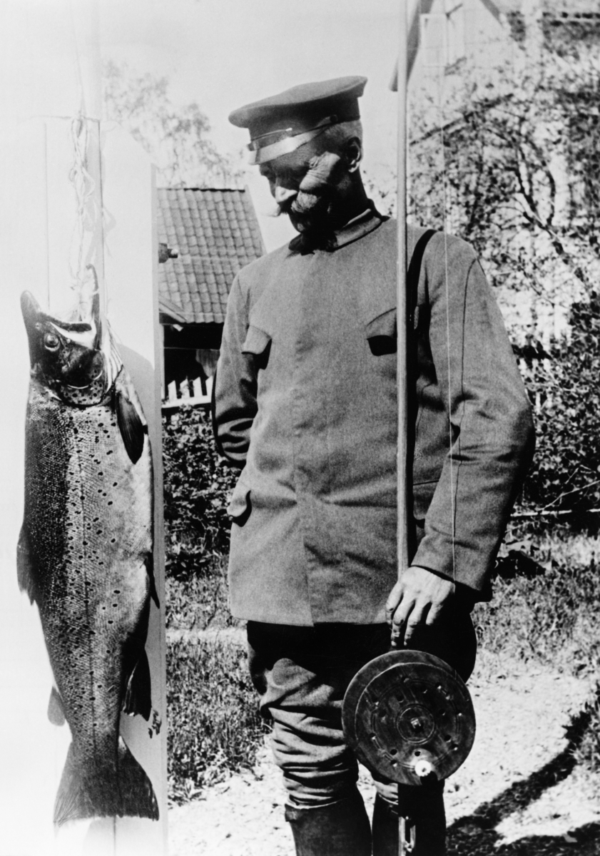 Hamarmannen Knut Emilsen (1876-1942), fotografert ved en 13 kilo tung og 90 centimeter lang ørret han tok i Mjøsa utenfor Snekkersvea på Nes (på vestsida av Furnesfjorden) den 2. juni 1925.  Emilsen hadde hengt storfisken i flaggstanga.  Sjøl sto han ved siden av, kledd i ei uniformsliknende jakke uten distinksjoner og med ei lue med blank skygge på hodet.  Med venstre hånd støttet han et «dreggsprøtt» - ei fiskestang med ei diger tresnelle, en redskapstype som ble brukt under dorging i Mjøsa.  Fotografiet er tydeligvis tatt i fiskerens egen hage.  Vi skimter stakittet mot nærmeste gate og bebyggelsen på motsatt side.  Knut Emilsen var født på en av husmannsplassene under garden Stor-Imerslund i Vang, men som voksen bodde han i Grønnegata på Hamar.  Han var bryggeriarbeider av yrke.
Mjøsørret. Mjøs-ørret. Hunderørret. Dreggeprøtt. Fiske. Sportsfisker. Sportsfiske. Dorging. Fangst.