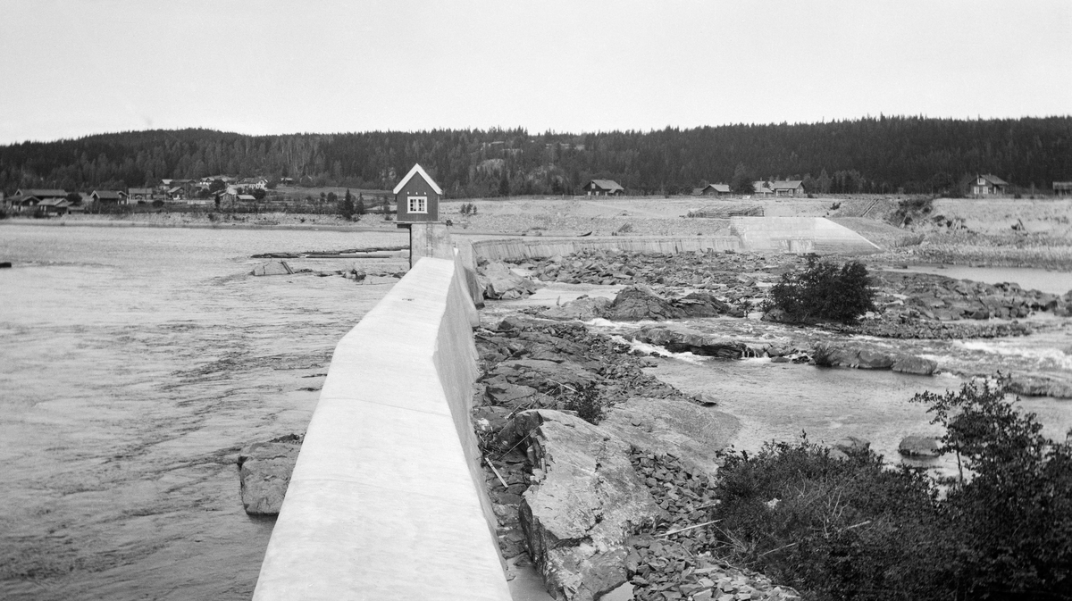 Damanlegget ved Skjefstadfossen i Glomma.  Skjefstadfossen ligger i Heradsbygda i Elverum, og det første kraftanlegget her ble reist i 1909-1910.  Dette fotografiet er antakelig tatt like etter at dammen var ferdig, sannsynligvis i 1910.  Fotografen har stått ved den vestre enden av krona på den betongstøpte dammen, med kameraet vendt østover.  Vi ser dammen og med et lite lukehus, uført i bordkledd bindingsverk og med forholdsvis spisst saltak.  Ved den østre elvebredden ender dammen i et kraftig, betongstøpt landkar.  På elvebrinken bakenfor skimter vi bebyggelsen på flere små gardsbruk, foran en lav åsrygg som er kledd med barskog. 

Litt mer informasjon om kraftanlegget ved Skjefstadfossen finnes under fanen «Andre opplysninger». 