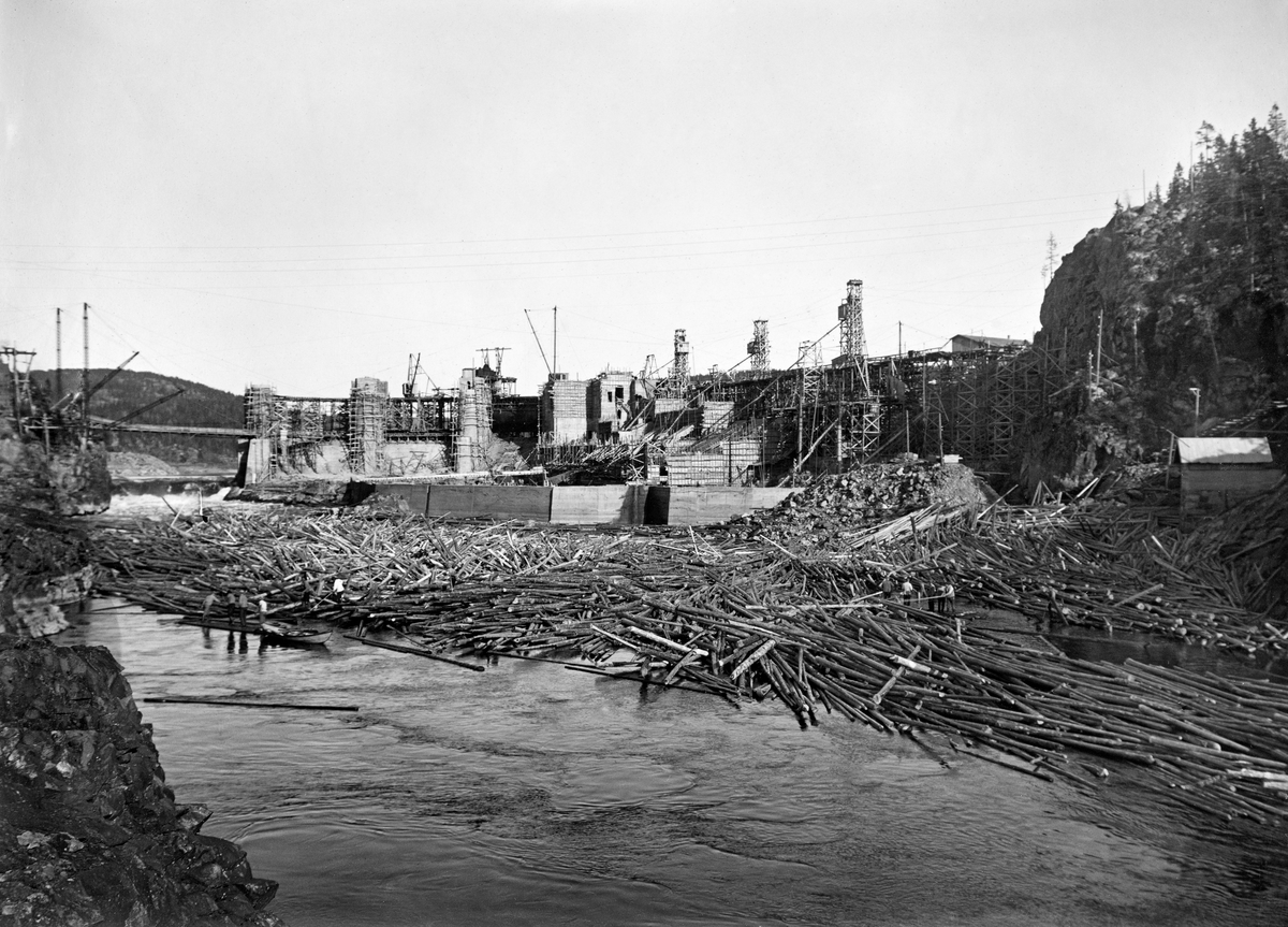 Solbergfossen i Askim, fotografert i forbindelse med bygginga av en dam og et kraftverk ved denne fossen.  Fotografiet er tatt nedenfra (i motstrøms retning).  Sentralt i bildet ser vi forskalinger til betongkonstruksjoner.  En betongskjerm langs elvebredden foran tomta til kraftverksbygningen var allerede ferdigstøpt.  Foran denne skjermen hadde det bygd seg opp en betydelig tømmervase (haug).  I bakgrunnen ser vi hvordan vannet fortsatt fikk strømme fritt gjennom den vestre delen av damløpet, mens bygge arbeidet gikk sin gang lengre øst.  Dette fotografiet ble tatt i 1921.  Kraftverket ble satt i drift tre år seinere.