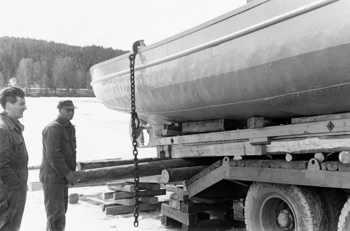 Transport av varpebåten «Varp I» fra Fetsund til Ullern ferjested i Sør-Odal, Hedmark.  Fotografiet er tatt fra sida og viser akterenden av Varp I og den bakre delen av bilen.  Båten ligger bakpå på et plan som antakelig ellers ble brukt til transport av anleggsmaskiner.  Fotografiet viser hvordan båten var støttet opp på ei treramme med profil avpasset etter skrogets ytre krumning.  Poenget med denne ramma må primært ha vært å forebygge at Varp I skulle bikke på kjølen under transport på svingete veger.  Fotografiet illustrerer også hvordan fartøyet skulle trilles på et skråplan med tversoverliggende tømmerstokker ned mot vassdraget. To karer står til venstre på bildet og betrakter situasjonen.  Spedisjonsfirmaet P. Nielsen fra Oslo som hadde ansvar for transporten. 

«Varp I» eller «VB 51» ble bygd for Christiania Tømmerdirektion i 1914.  Båten ble i hovedsak bruk på Glomma i nedre Solør og framfor alt på Storsjøen i Odalen.  Den hadde sitt opplagssted på Bukkeneset i Nord-Odal.  Der ble det opprinnelige overbygget, motoren og vinsjen fjernet like før jul i 1954 (jfr. SJF.1990-02242 og SJF.1990-02243).  Så ble skroget kjørt til Glomma fellesfløtingsforenings lenseanlegg ved Fetsund i Akershus, hvor båten ble ombygd og modernisert, før den altså ble sjøsatt ved Ullern ferjested påfølgende vår for fortsatt bruk i fløtinga i den nevnte regionen.