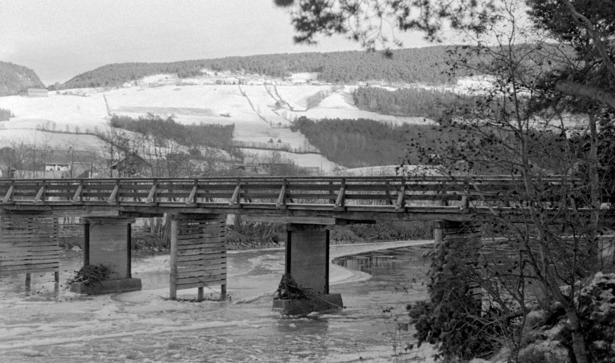 Bru over Gudbrandsdalslågen ved tettstedet Dovre i kommunen med samme navn.  Fotografiet ble tatt en kald oktoberdag i 1955.  Vannstanden i elva var lav, og det var i ferd med å danne seg ei isskorpe på vannflata.  Brua var en bjelkekonstruksjon av tre, der vegbanen kviler på stolper som er plassert på betongfundamenter i elveløpet.  I bakgrunnen til venstre skimter vi noe av bebyggelse vest for Dovre jernbanestasjon.  I bakgrunnen ser vi Åsgrenda med snødekte jorder, og til venstre skimter vi skaret der Dragåa renner ned mot hoveddalføret og Lågen.