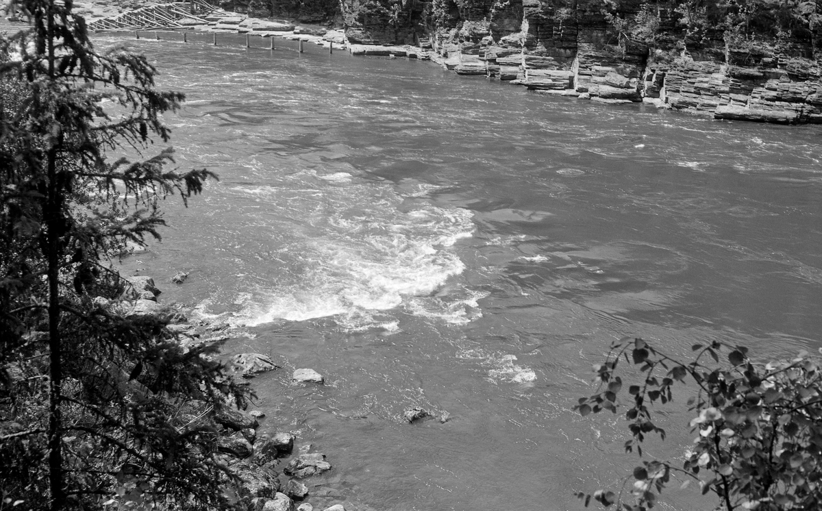 Elveparti fra Gudbrandsdalslågen, nedenfor Hunderfossen.  Fotografiet er tatt fra en bratt elveskrent på Øyer-sida av elva, ned mot et vannspeil der Lågen går forholdsvis strid over steinete grunn mellom to berg.  Øverst til venastre i bildet ser skimter vi et parti langs elvebredden som var pælet.  Fotografiet ble tatt i 1961, antakelig for å dokumentere vannføringa før kraftutbygginga i den ovenforliggende Hunderfossen ble ferdigstilt.