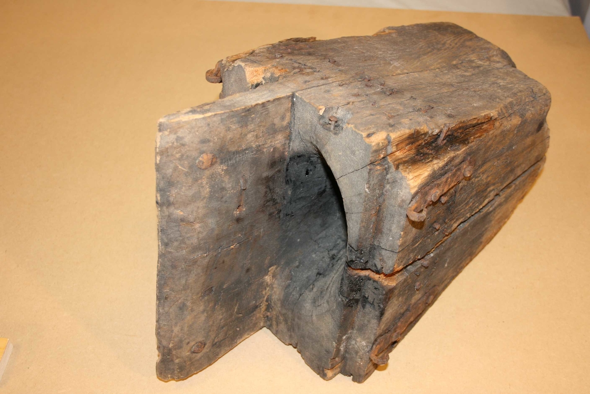 Firkanta trestykke med røyrforma hol gjennom. Mange spikrar, restar av beslag. Sprekt, delvis roten, brannskader ytst.