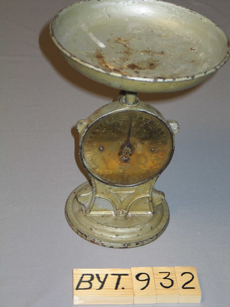 Rund fotplate, metall. Klokkeforma talskive med vektskala i gram og kilo, med visar. Sjølve talskiva av messing. Skål på toppen. Grågrøn måling.