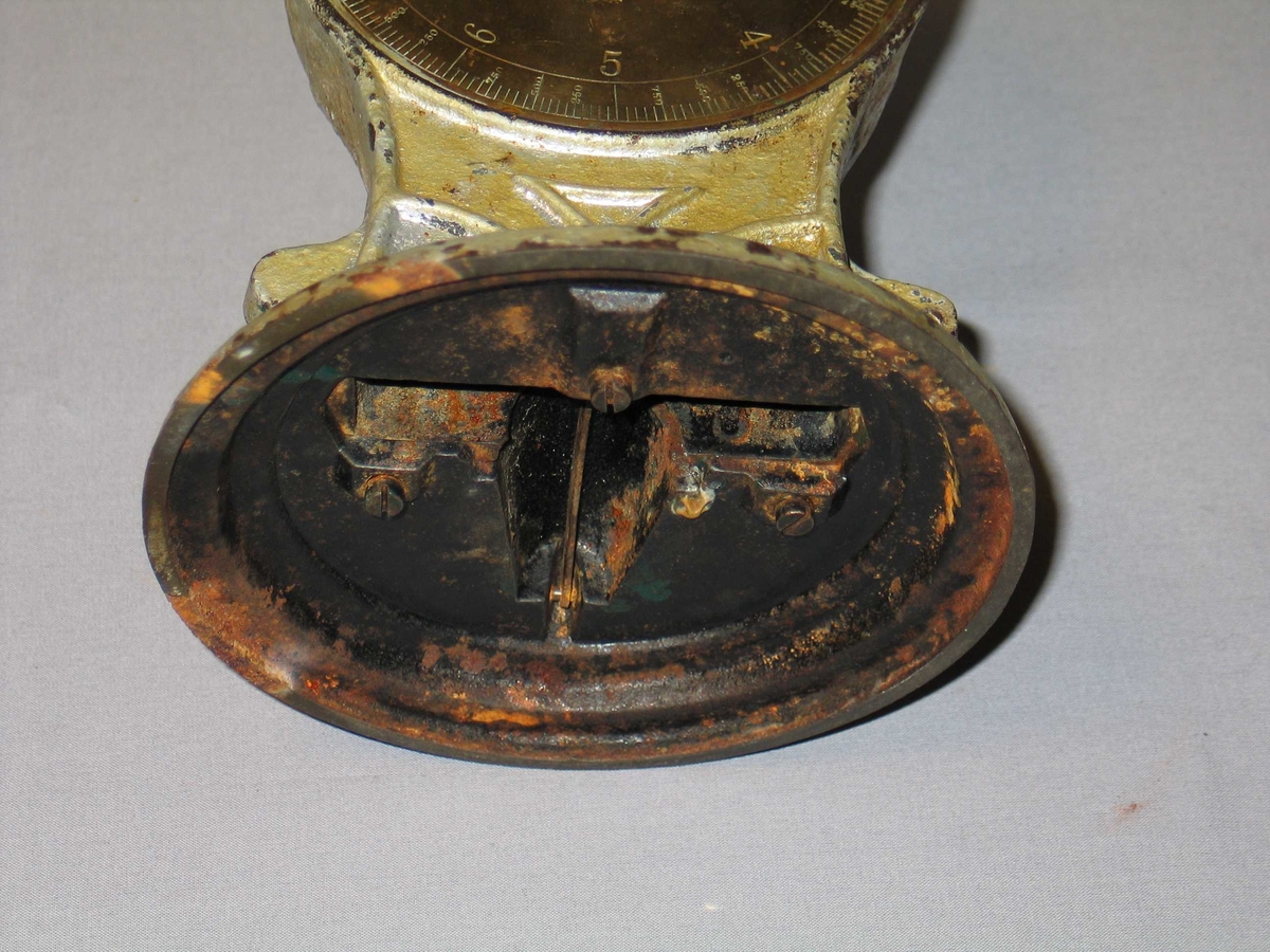 Rund fotplate, metall. Klokkeforma talskive med vektskala i gram og kilo, med visar. Sjølve talskiva av messing. Skål på toppen. Grågrøn måling.