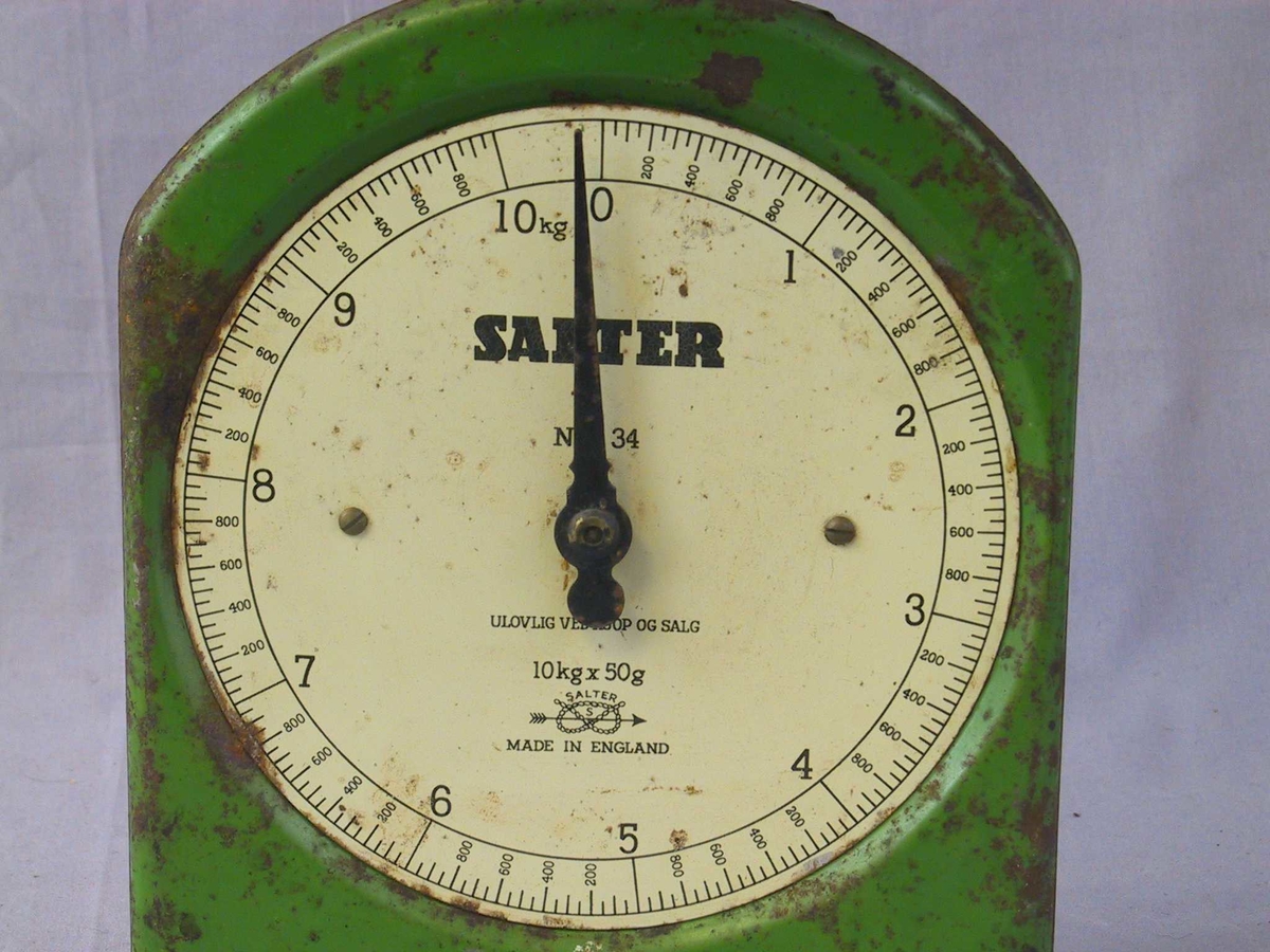 Flat framside med rett botn og rette sider, avrunda topp. Kvit, klokkeforma talskive med vektskala og visar. Laus skål som står på kryssforma, flate berepinnar på toppen av stav som går ned i halvsylinderforma bakstykke. Grønmåla med kvit talskive og svarte tal.