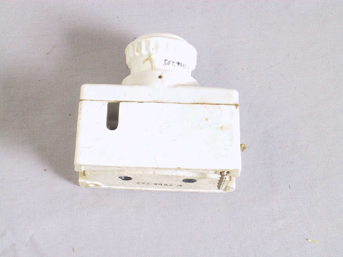 Rektangulær boks av kvit porselen. Sikringshylse med sikring skruva i opninga midt oppå