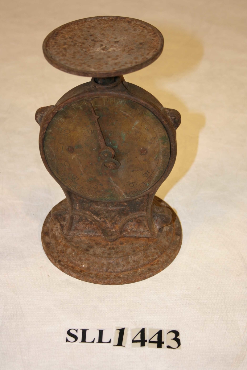 Rund fotplate, metall. Klokkeforma talskive med vektskala i gram og kilo, med visar. Sjølve talskiva av messing. Rund plate på toppen, manglar skål. Rusta.