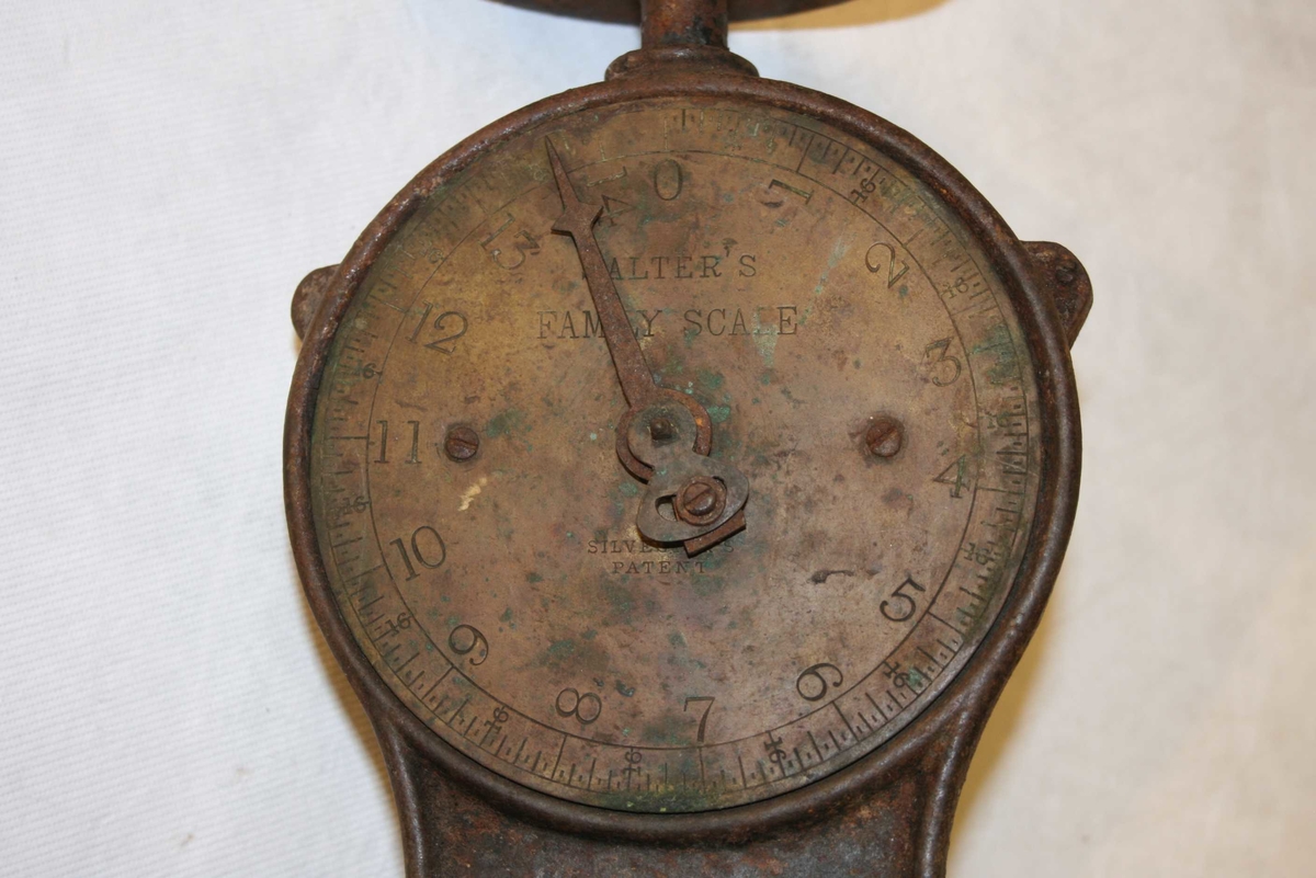 Rund fotplate, metall. Klokkeforma talskive med vektskala i gram og kilo, med visar. Sjølve talskiva av messing. Rund plate på toppen, manglar skål. Rusta.