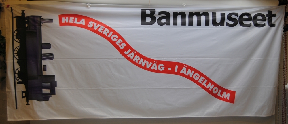 Vit flagga med dekoration av ånglok och text.
Längst upp står det "Banmuseet" i svart. Nedanför detta står det "HELA SVERIGES JÄRNVÄG - I ÄNGELHOLM" med vit text i ett rött böljande språkband. Utmed vänster kortsida är det ett svart ånglok.
Flaggan har plastöglor utmed den övre långsidan.