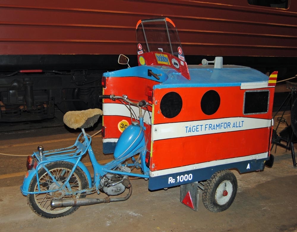 Trehjulig moped som är omgjord för att likna ett Rc-lok. Skåpet är målat orange med svarta fält som föreställer fönster, turkos tak, ett vitt fält med svarta kantlinjer mitt på med texten "TÅGET FRAMFÖR ALLT" i turkos och en vit och en blågrå kant nedtill där det står "A" och "Rc 1000" med vita bokstäver. I takets främre hörn sitter rödgul randiga skärmar. Mitt fram sitter en grå och röd tuta. På taket sitter en vindruta av plast med flera klisterdekaler på. Dekalerna på framsidan av vindrutan är symmetriskt uppsatta på var sida och föreställer TV4s symbol "4", en blixt som är symbol för elektricitet och dekaler med texten "allt om hobby". På insidan sitter också reklam dekaler från TV4, Allt om hobby samt en dekal med ett ånglok från TGOJ och en dekal från Sundbyberg. På skåpet, till vänster framför mopeden, sitter en dekal som föreställer en hastighetsskylt för 30 km/tim. Taket går att öppna som en lucka och har öglor för hänglås på vänster sida. På vänster sida sitter en backspegel. Mopeden är turkos. Sadeln har ett skydd av fårskinn. Framtill sitter lyktor och buffertar, målade svarta fönster med konstgjorda vindrutetorkare, en SJ klisterdekal i mitten och "1000" målat på sidorna. Plog finns i skåpet.