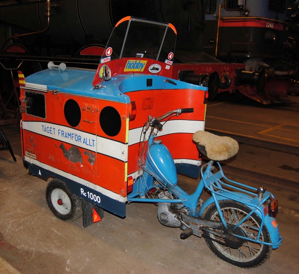 Trehjulig moped som är omgjord för att likna ett Rc-lok. Skåpet är målat orange med svarta fält som föreställer fönster, turkos tak, ett vitt fält med svarta kantlinjer mitt på med texten "TÅGET FRAMFÖR ALLT" i turkos och en vit och en blågrå kant nedtill där det står "A" och "Rc 1000" med vita bokstäver. I takets främre hörn sitter rödgul randiga skärmar. Mitt fram sitter en grå och röd tuta. På taket sitter en vindruta av plast med flera klisterdekaler på. Dekalerna på framsidan av vindrutan är symmetriskt uppsatta på var sida och föreställer TV4s symbol "4", en blixt som är symbol för elektricitet och dekaler med texten "allt om hobby". På insidan sitter också reklam dekaler från TV4, Allt om hobby samt en dekal med ett ånglok från TGOJ och en dekal från Sundbyberg. På skåpet, till vänster framför mopeden, sitter en dekal som föreställer en hastighetsskylt för 30 km/tim. Taket går att öppna som en lucka och har öglor för hänglås på vänster sida. På vänster sida sitter en backspegel. Mopeden är turkos. Sadeln har ett skydd av fårskinn. Framtill sitter lyktor och buffertar, målade svarta fönster med konstgjorda vindrutetorkare, en SJ klisterdekal i mitten och "1000" målat på sidorna. Plog finns i skåpet.