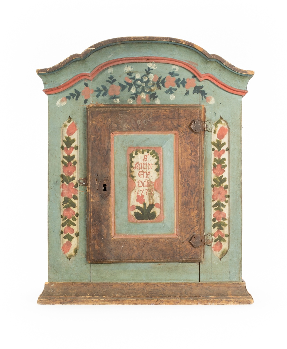 Hängskåp med svängt krön. Enkel lucka. Målat i blått med bruna sidor. En bred brun ådrad ram runt om luckan. Vegetativ ornering i grönt, rosa och blått. Märkt på luckan: "J Karin Erik Dåtter 1777". Skåpet är inuti omålat och med en hylla. Luckans insida vitmålad med blå ornering.